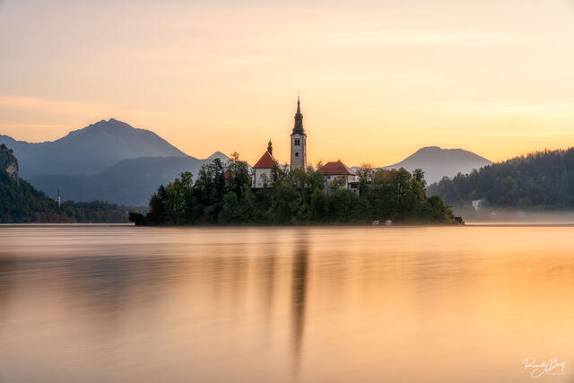 sunrise over the Lake Bled Monastery with mist and colorful hues in the sky.