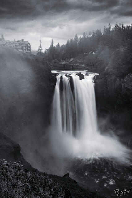 fine art photograph of Snoqualmie Falls, captured in black and white. The waterfall is seen from a distance, cascading down a rocky cliff into a pool of water.